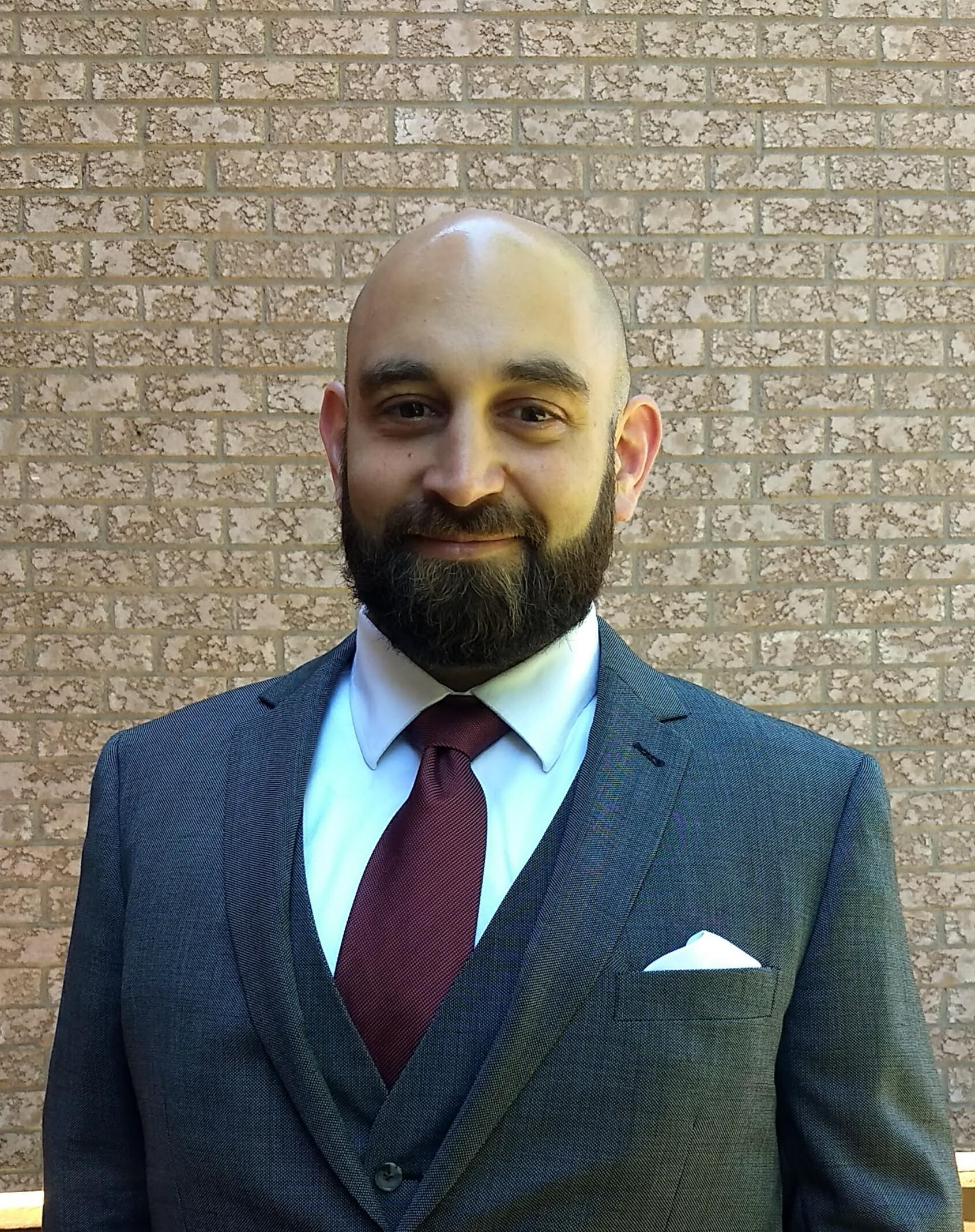 Smiling man in three piece grey suit against a brick wall backdrop.