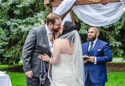 Happy couple kissing on completion of an outdoor wedding ceremony with their officiant in the background.